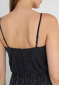 Vestido azul marino con tirantes finos, que presenta rayas metálicas verticales y un diseño plisado, resaltando un corpiño ajustado y una falda fluida.