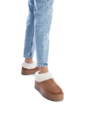 Zapatillas marrones con un suave collar de piel sintética blanca, con una suela gruesa y un sencillo detalle de costura, combinadas con unos vaqueros de color azul claro.