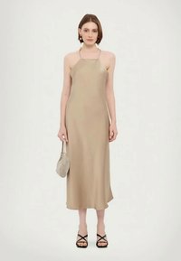 Sieviete ģērbjas bēša halter-stila midi kleitā, melnos stilu sandalos ar siksniņām, valkā zeltainas auskarus un rokās tur mazu bēša toņa rokassomu.