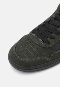 Dark green suede sneaker with black accents, featuring decorative stitching, perforations, and a textured rubber outsole.
