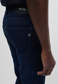 Närbild av person som bär mörkblå jeans med BOSS-logotyp på midjebandet och metallisk emblem på bakfickan.