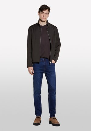 Un modello maschile si presenta con sicurezza indossando una giacca scura sopra una camicia nera, abbinata a jeans blu e scarpe marroni, su uno sfondo chiaro.