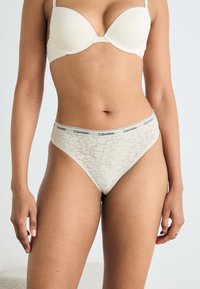 Spennende blonde tanga i lys beige med elastisk midjebånd som har svart "Calvin Klein" merkevare. Glatt tekstur, gjennomsiktige blonde mønsterdetaljer.