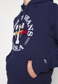 Námořnická modrá mikina s kapucí se bílým a červeným vyšitým logem, se stahovací kapucí a přední kapsou. Rukávy s branded pruhy na manžetách.