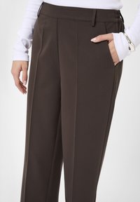 Braune, maßgeschneiderte Hose mit glatter Textur, Bundfalten und seitlichen Taschen. Tragen mit einem weißen Langarmoberteil. Mit Gürtelschlaufen ausgestattet.