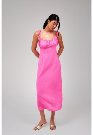 Jonge vrouw in een felroze midi-jurk met bindbandjes, staand met de handen achter de rug, naar beneden kijkend, met gouden open-teen hakken.