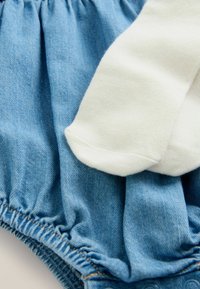 Denim romper dengan pinggang yang dihimpun, warna biru muda, dan kaus kaki katun putih dengan ujung bulat, terletak datar di atas kain.