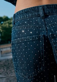 Tumed sinised teksapüksid, millel on väiksed peegeldavad nööbid tagataskul ja üla-piha piirkonnas, looduskaunis väliskeskkonnas.