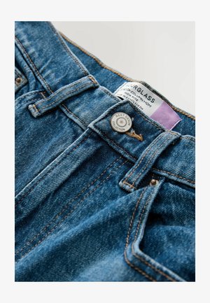 Magas derekú, kék mosott farmernadrág, feltűnő varrással, fémből készült gombzárral, és a "NEXT DENIM COLLECTION" felirattal ellátott címkével.