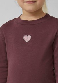 Bordeauxfarbendes Langarmshirt mit einem pinken Glitzerherz-Patch mit einem Riss, auf einem glatten Baumwollstoff.