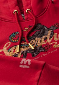 Sudadera roja con ojales y cordones dorados, que presenta un diseño gráfico texturizado en multicolores y un logo sutil en la manga.