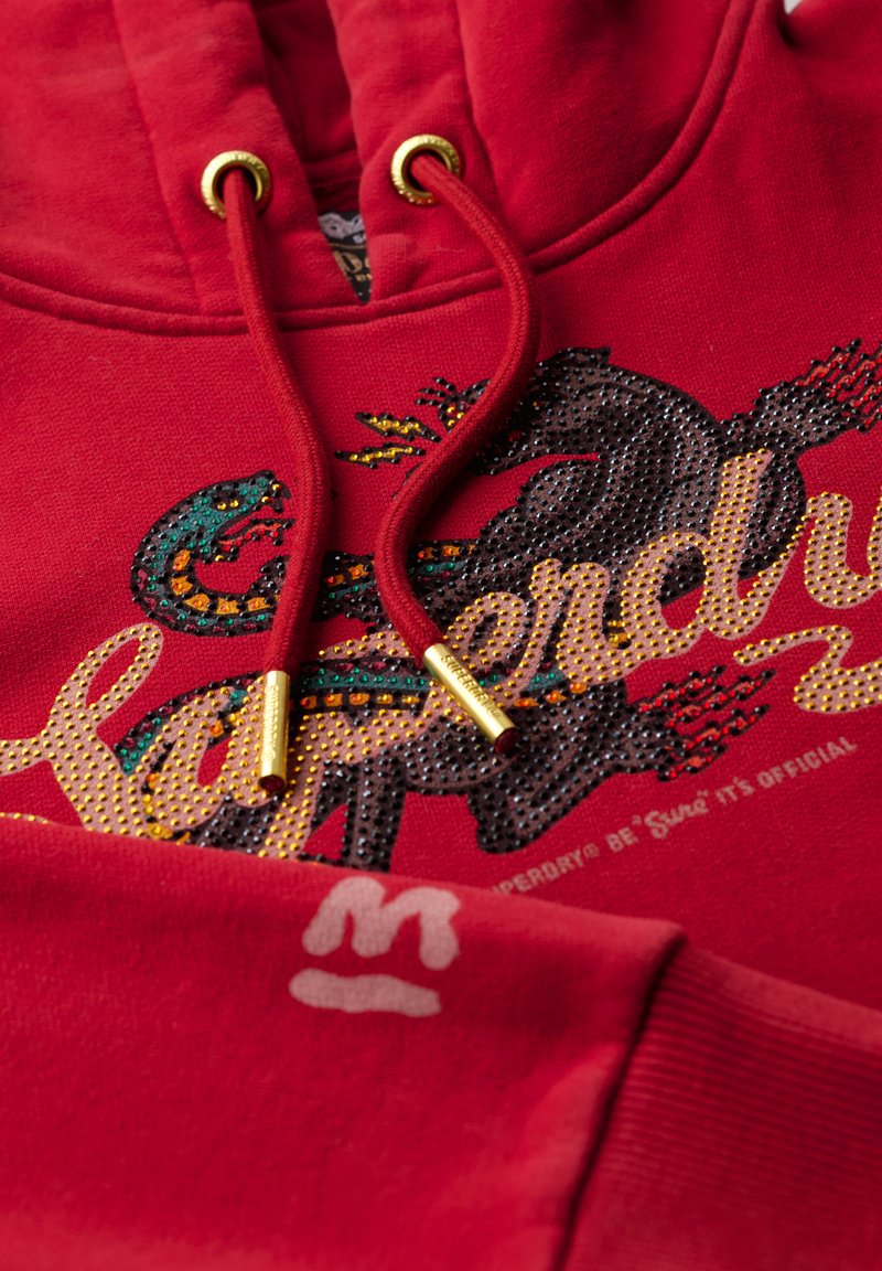 Czerwona bluza z kapturem z złotymi oczkami i sznurkami, z teksturowanym wzorem graficznym w wielu kolorach oraz subtelnym logo na rękawie.