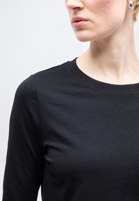 Κοντινό πλάνο ενός ατόμου που φοράει μαύρο μπλουζάκι με στρογγυλή λαιμόκοψη και ασημένιο κρίκο στο αυτί, με ουδέτερη έκφραση προσώπου, στραμμένο προς τα δεξιά.