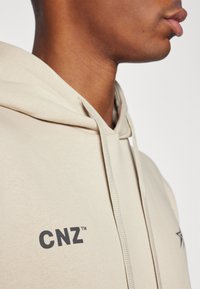 Felpa beige con cappuccio e cordino; presenta un logo frontale "CNZ" in nero. Tessuto liscio con una morbida texture, che mette in mostra un design casual.
