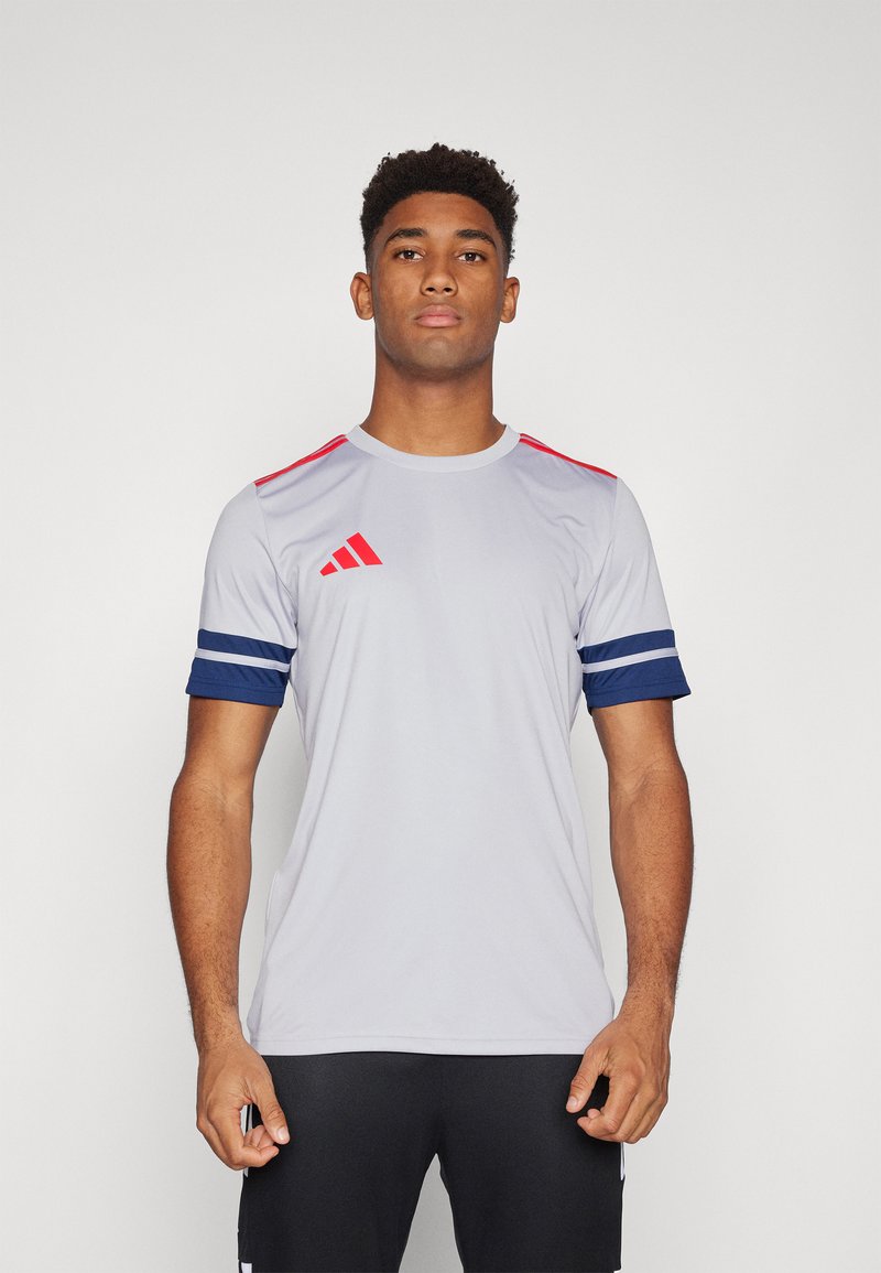 Camiseta atlética gris de manga corta, con detalles rojos en los hombros y una franja azul marino en los puños. La parte delantera muestra el logo de tres rayas.