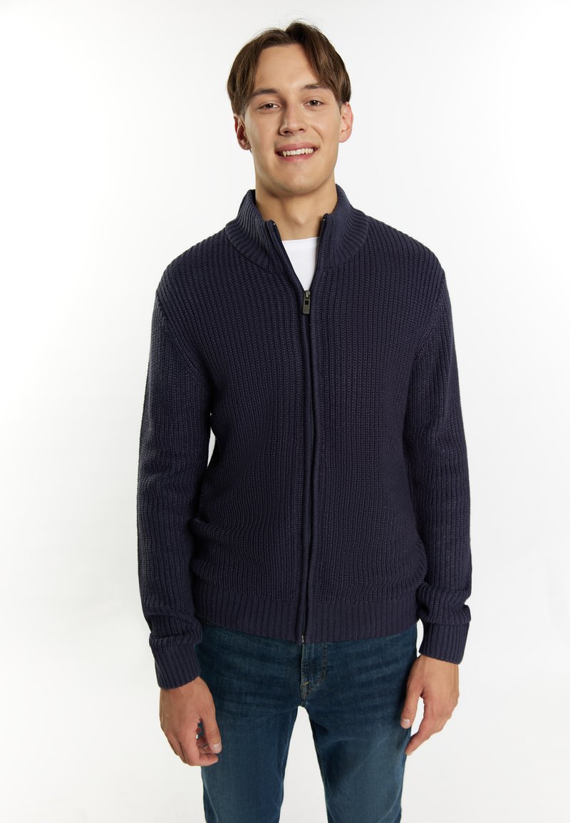 Maglione lavorato a maglia di colore blu navy con colletto alto e zip frontale; presenta una texture a coste e maniche lunghe; indossato sopra una camicia bianca.