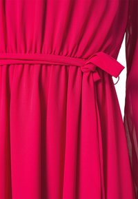 Robe rose vif avec taille froncée et ceinture en tissu nouée en nœud sur le côté, présentant une longue manche en tulle partiellement visible.