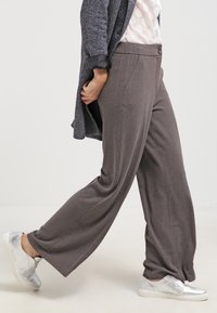Pantalon gris à jambes larges avec une texture lisse et une légère brillance, doté de poches avant et d'une fermeture à bouton. Associé à des baskets argentées.