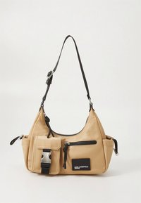 Beige skulderveske i nylon med svart justerbar stropp, glidelåslommer og en frontlomme med sølvspenne.