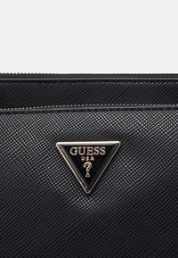 Musta tekstuuriga kott kuldse tsipi ja kolmnurkse logoga, millel on kiri "GUESS USA" ja küsimärgi sümbol keskel.