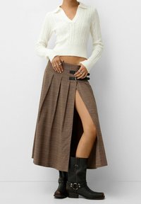 Brązowa spódnica w kratę z bocznym rozcięciem, z wysokim stanem i czarnymi elementami ozdobnymi. Połączona z białym swetrem w warkocze i czarnymi butami.