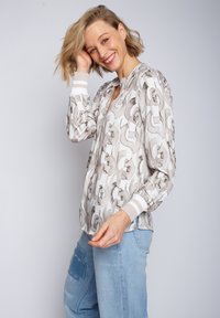 Leichte Bluse mit beigem und grauem Kettenmuster, mit V-Ausschnitt und gerippten Bündchen; kombiniert mit blauen Jeanshosen.