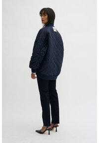 Quilted navy bomber jacket with a high collar and ribbed cuffs, paired with dark slim jeans and black high-heeled shoes. Tag visible on back.