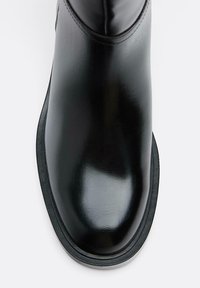 Črni gležnar iz usnja z sijočim zaključkom, zaobljenim sprednjim delom in kontrastnim gumijastim podplatom. Ima minimalno šivanje in eleganten dizajn.