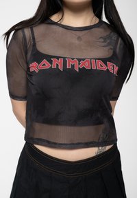 Black mesh crop top featuring a prominent red "IRON MAIDEN" graphic, short sleeves, and a sheer texture. Styled with a black skirt.