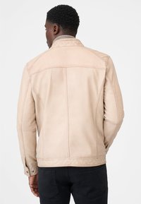 Mand står med ryggen til iført en lys beige læderjakke og sorte bukser foran en almindelig hvid baggrund.