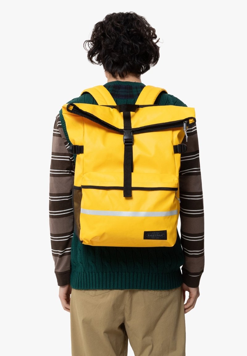Personne portant un pull vert et une chemise à manches longues rayée avec un sac à dos jaune à fermeture roulante, avec des bretelles noires et une bande réfléchissante dans le dos.