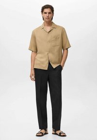 Camicia a maniche corte beige con bottoni e tasca sul petto, abbinata a pantaloni neri e sandali neri. Tessuto morbido e vestibilità comoda.