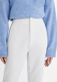 White tailored pants with vertical pleats paired with a light blue knit sweater, highlighting texture and fit. Soft, relaxed silhouette.