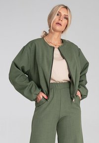 Zielona bomberka z zamkiem z przodu, szerokimi rękawami i teksturowanym wykończeniem, w zestawieniu z pasującymi zielonymi spodniami z wysokim stanem i beżową bluzką.