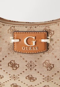 Gros plan sur un tissu beige à motifs avec un patch en cuir marron affichant le logo argenté "G" et "GUESS", cousu au-dessus d'une fermeture éclair.