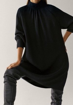 Femme portant une robe noire ample à manches longues avec un col montant, associée à des bottes en cuir noires au-dessus du genou, posant avec une jambe levée.
