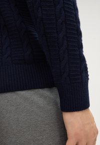 Maglione blu navy a maglia rasata con polsini a costine, caratterizzato da motivi testurizzati e una finitura morbida. Indossato sopra un tessuto grigio per creare un contrasto.