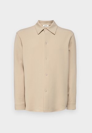 Beige Langarmhemd mit Knopfleiste, spitzem Kragen und silbernen Druckknöpfen, vor einem schlichten weißen Hintergrund präsentiert.