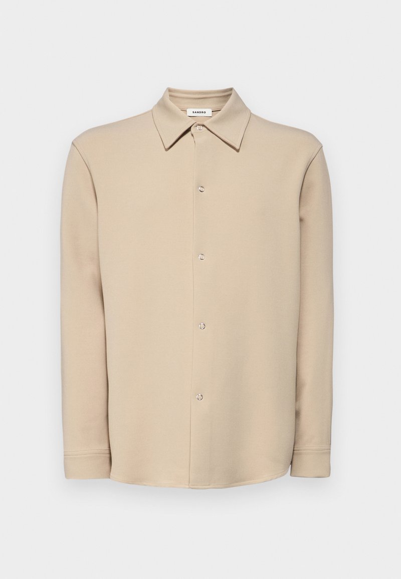 Beige overhemd met lange mouwen en knoopsluitingen, met een puntige kraag en zilveren drukknoopjes, weergegeven tegen een effen witte achtergrond.