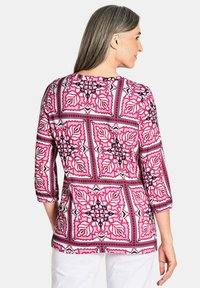 Roze top met driekwartmouwen, voorzien van een geometrisch bloemenpatroon met zwarte omtrekken en accenten. Gemaakt van een zachte, gladde stof.