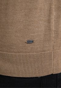 Pull en maille beige avec ourlet côtelé et petite étiquette ovale en métal portant le logo "Petrol" près du bord inférieur.