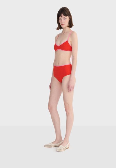 Femme debout de profil portant un bikini rouge avec des bordures roses et des chaussures tissées blanches à enfiler, sur un fond clair uni.