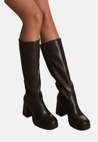 Jambes portant des bottes en cuir marron foncé hauteur genou avec des talons carrés et des zips latéraux, sur un fond clair uni.