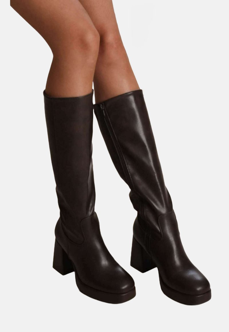 Jambes portant des bottes en cuir marron foncé hauteur genou avec des talons carrés et des zips latéraux, sur un fond clair uni.