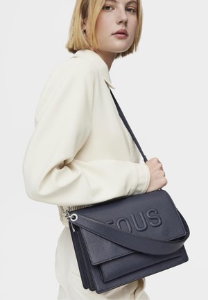 Borsa a tracolla in pelle blu con logo "TOUS" impresso, dotata di tre scomparti sovrapposti e una lunga tracolla regolabile.