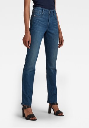 Woman wearing slim-fit blue jeans and black high-heeled sandals, standing against a plain white background.