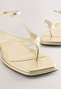 Sandali bassi dorati con design a T-strap, realizzati in materiale metallico. Punta quadrata, texture liscia e costruzione minimalista.