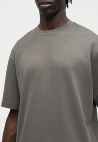 Άνδρας που φοράει απλό γκρι φαρδύ κοντομάνικο μπλουζάκι, με λαιμόκοψη και υφή υφάσματος.