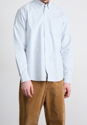 Mann trägt ein hellblaues langärmliges Hemd mit Knöpfen und braune Cordhosen, steht vor einem schlichten weißen Hintergrund.