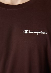 T-shirt en coton marron avec un col rond, arborant un logo "Champion" blanc sur le côté gauche. Le tissu semble lisse et doux.